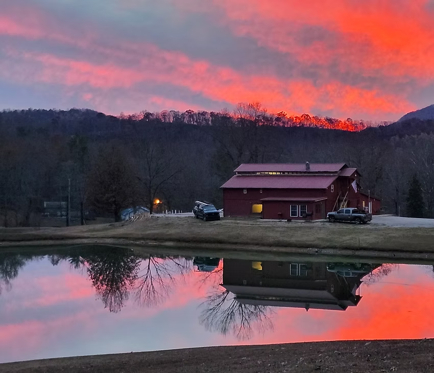 Red barn by a calm lake at sunset, with vivid pink clouds reflected in the water