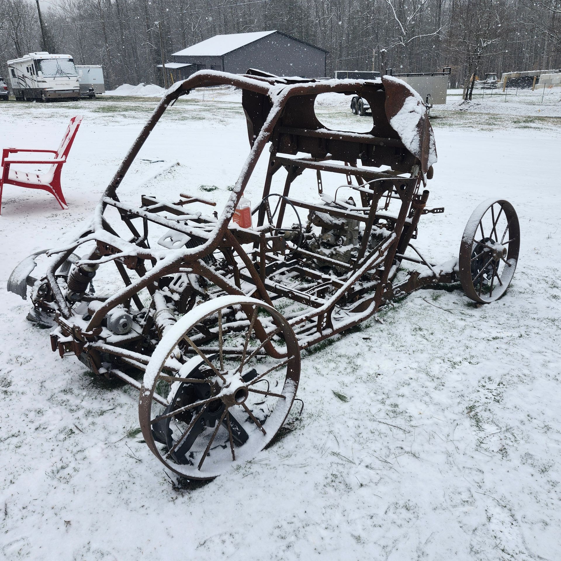 Snow-covered rusted metal go-kart frame in a yard, with small wheels and no body.