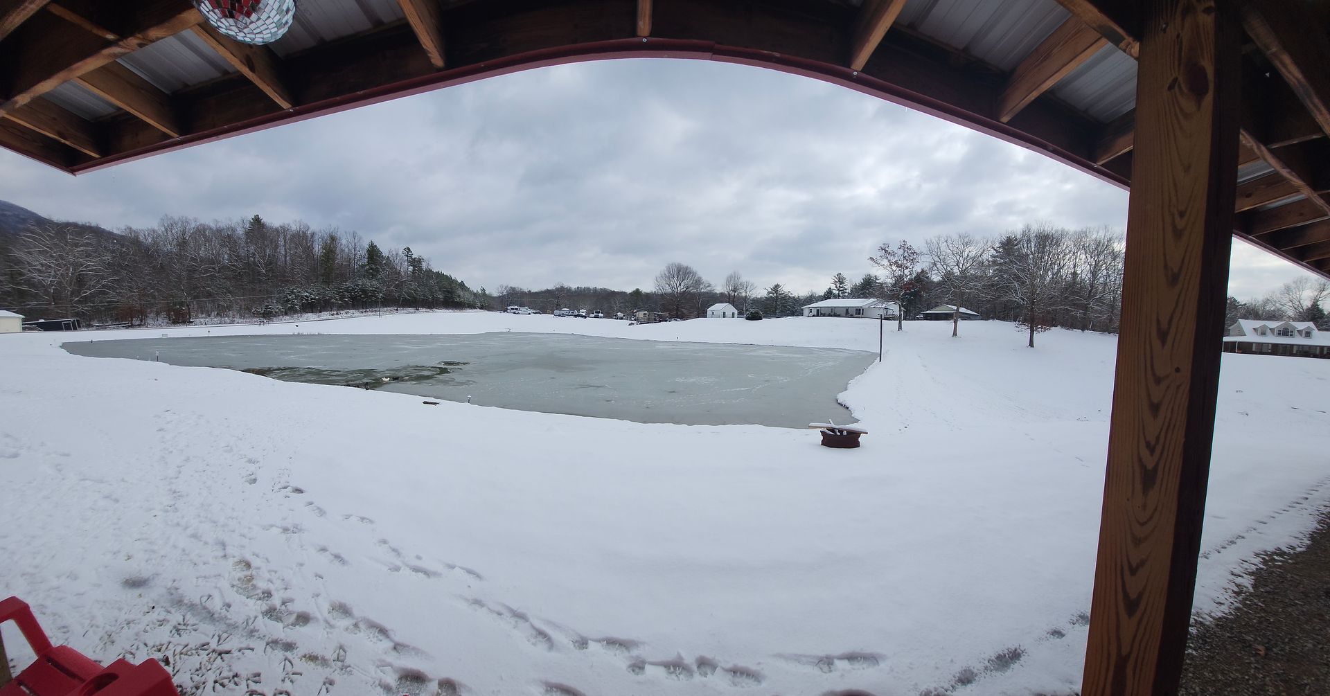 Snowy lakeside under a roofed pavilion, with a partially frozen lake and tree-lined shore.