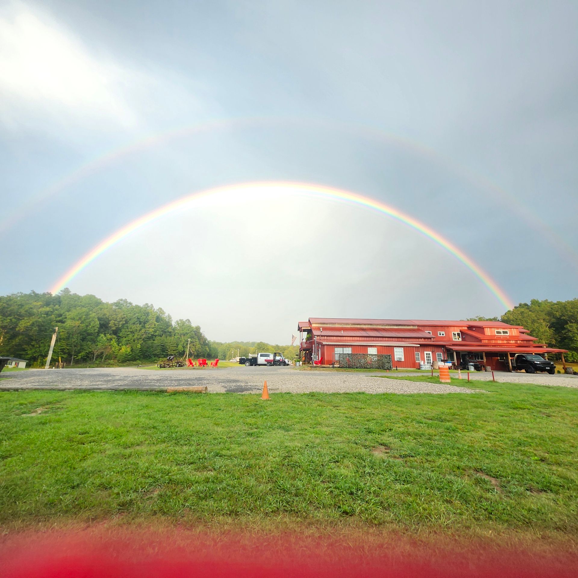 Rainbow over a grassy roadside with a red-roofed building and trees under a bright sky
