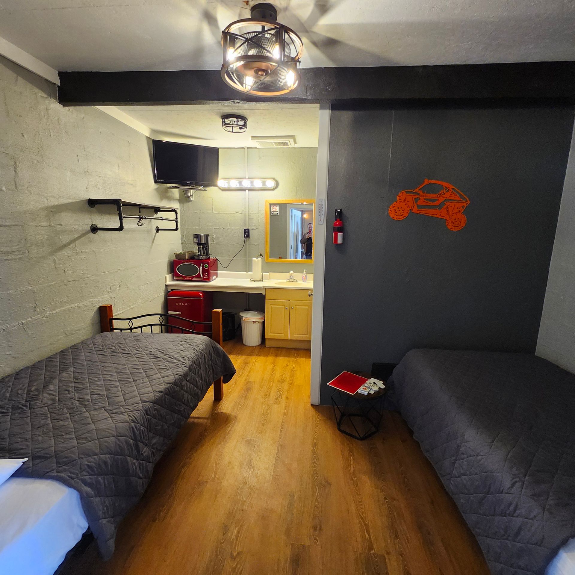 Small bedroom with two beds, wooden floor, desk, TV, and kitchenette nook under a ceiling light.