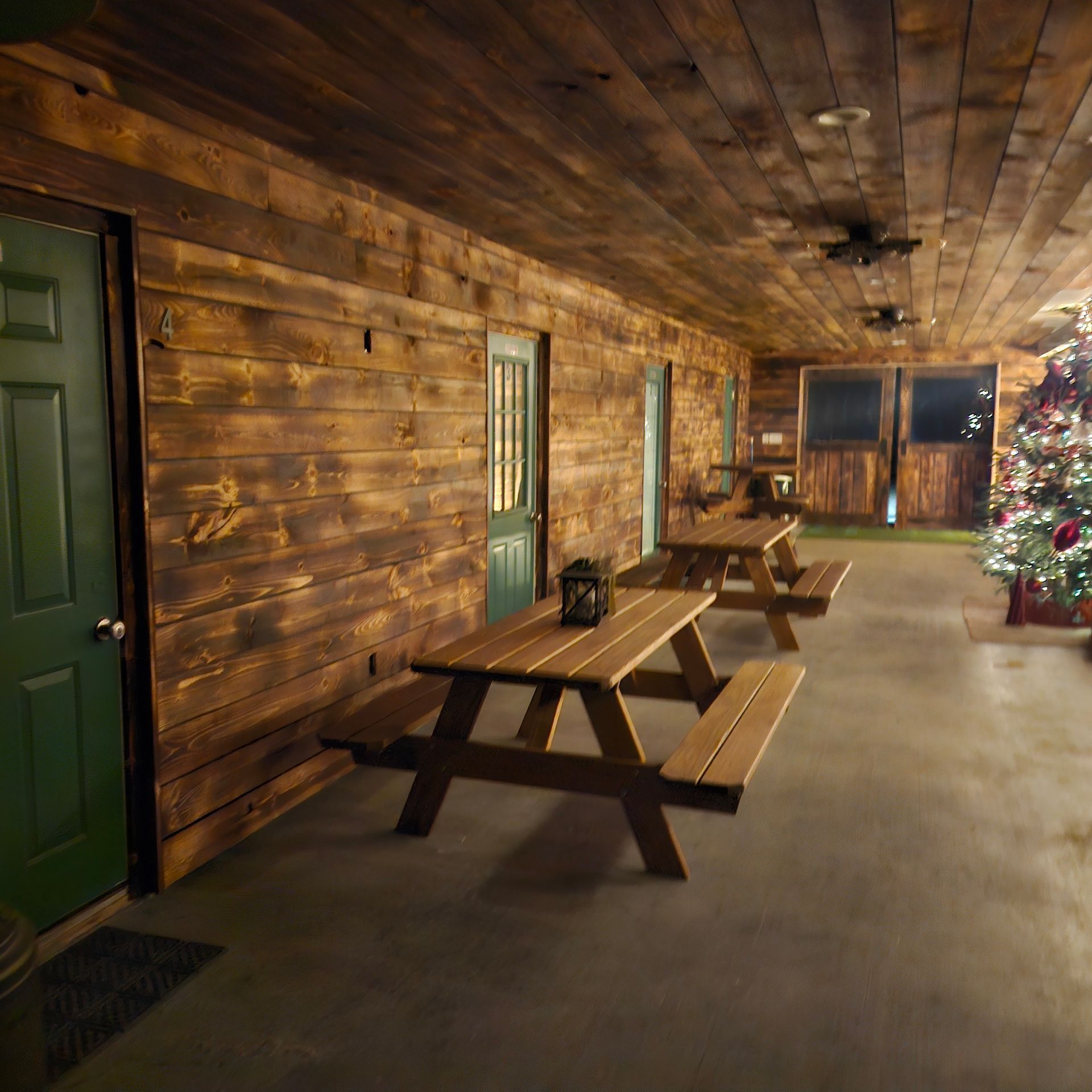 Covered wooden porch with picnic tables, green doors, and holiday lights in a rustic lodge-style setting