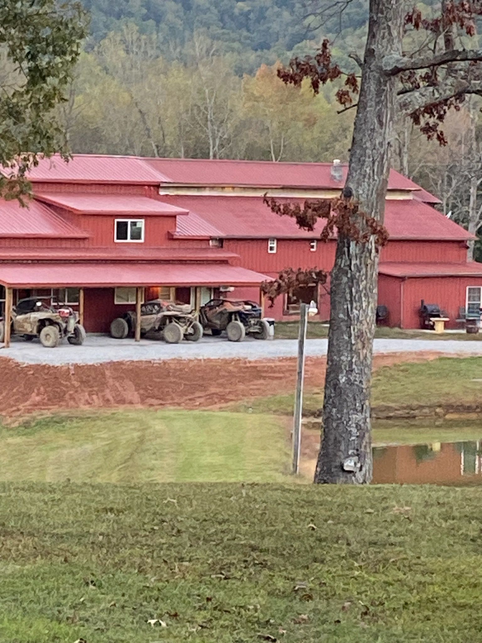 Red barn with red roof, tractors parked under the overhang, beside a grassy pond and wooded hillside.