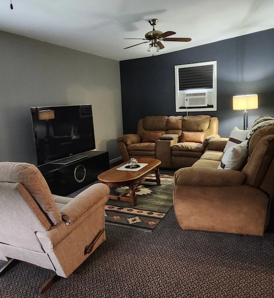 Cozy living room with tan recliners and sofa, dark TV stand, window AC, lamp, and ceiling fan.