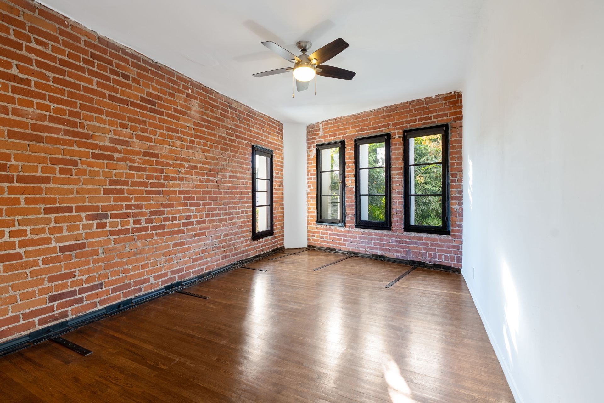 Room with exposed brick wall, three black-framed windows, hardwood floor, and ceiling fan.