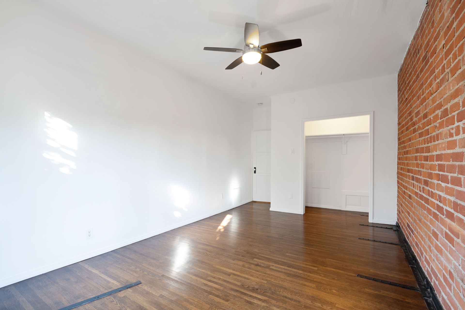 Empty room with hardwood floors, exposed brick wall, white walls, and a ceiling fan.
