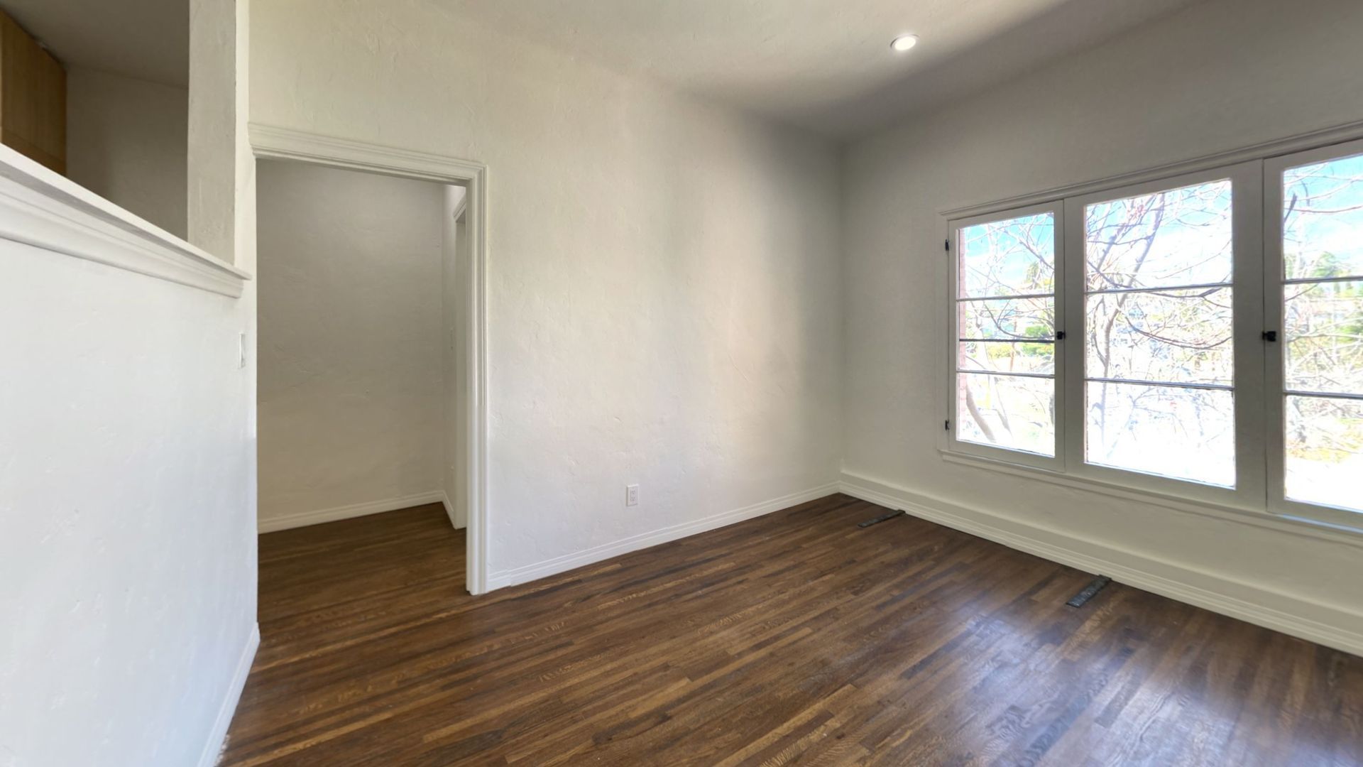 Empty room with hardwood floors, white walls, and a large window. An open doorway leads to another room.