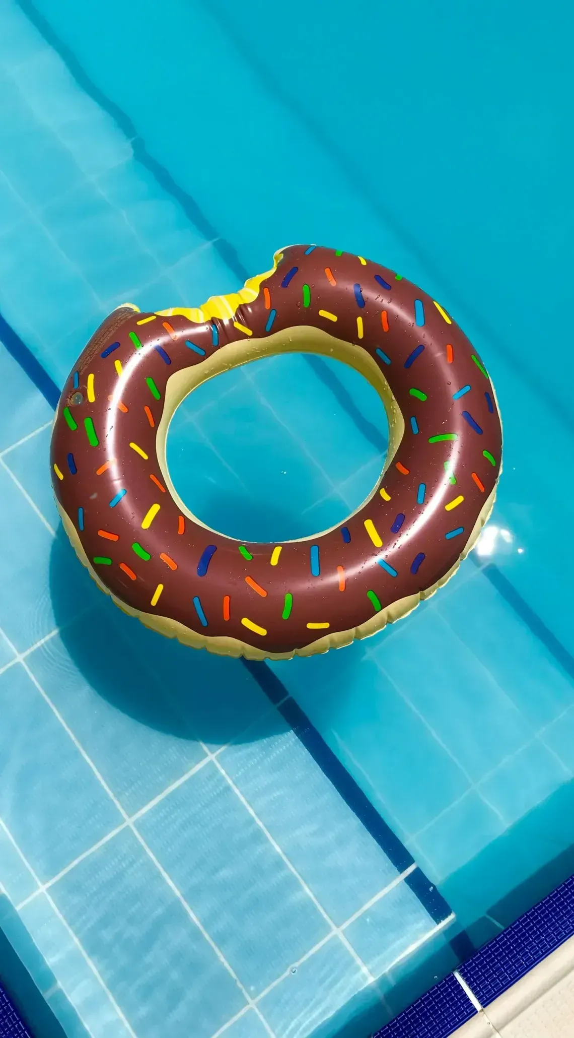 A chocolate donut-shaped pool float with colorful sprinkles floating on the blue water of a tiled swimming pool.