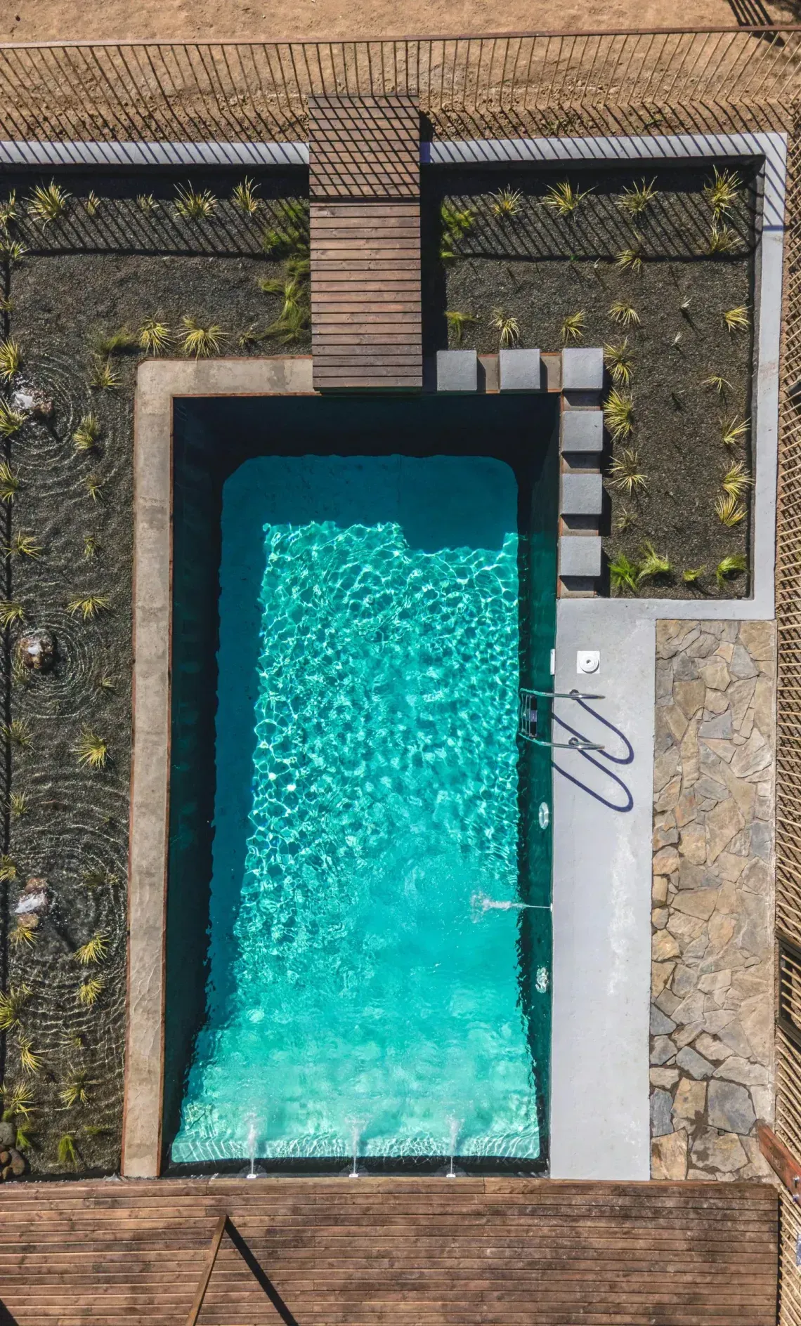 An aerial view of a rectangular, bright blue pool surrounded by dark gravel, a stone patio, and a wooden deck.