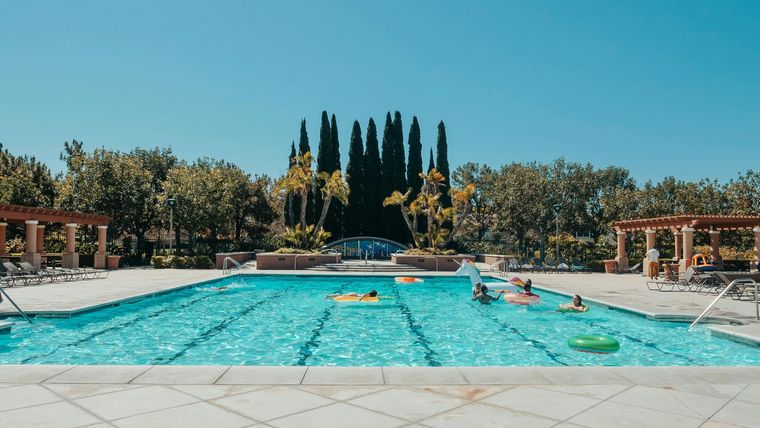 People relax and swim in a bright turquoise outdoor lap pool surrounded by trees and decorative structures.