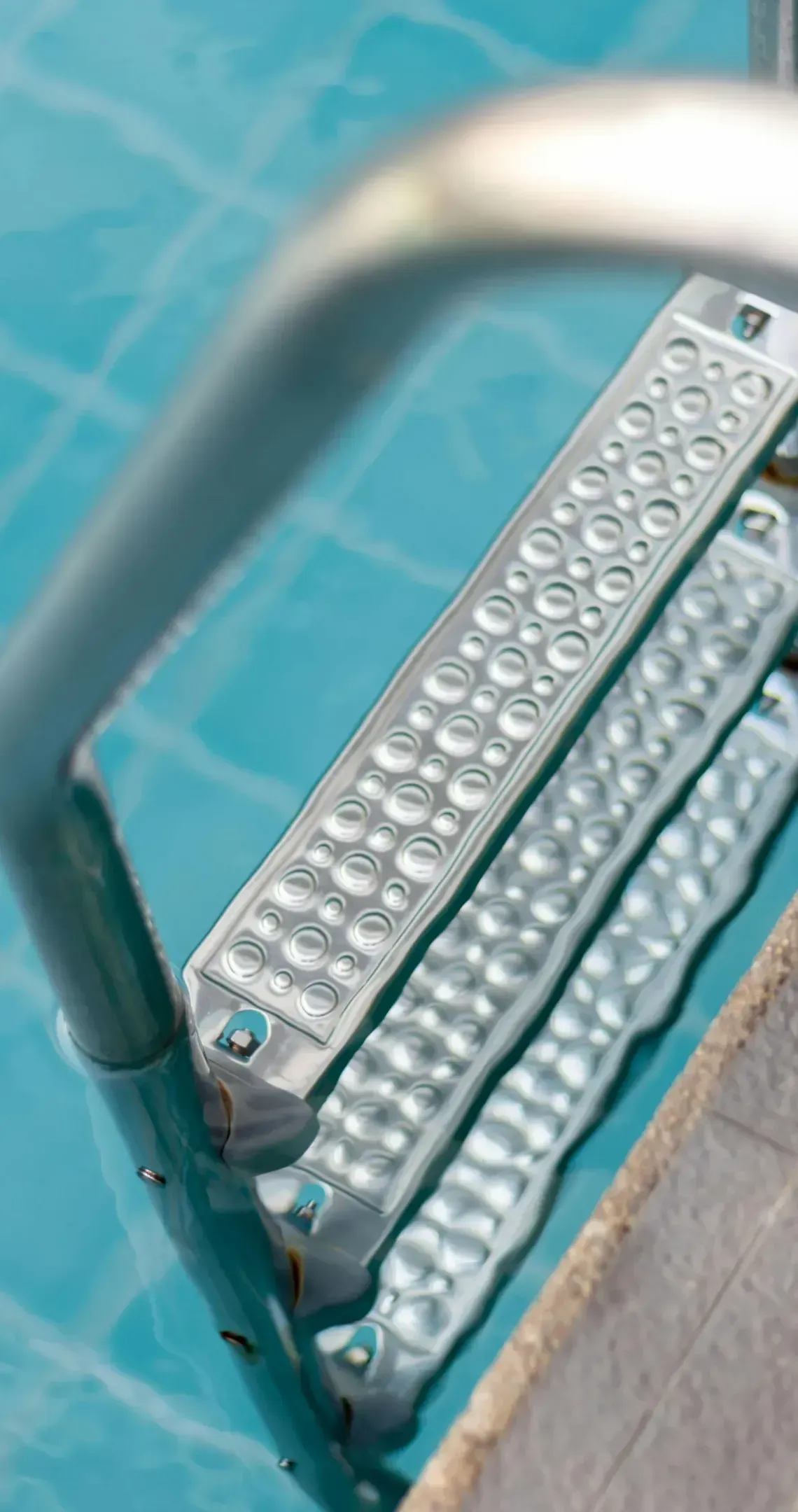 A metal pool ladder with textured steps submerged in light blue water next to a stone pool deck.