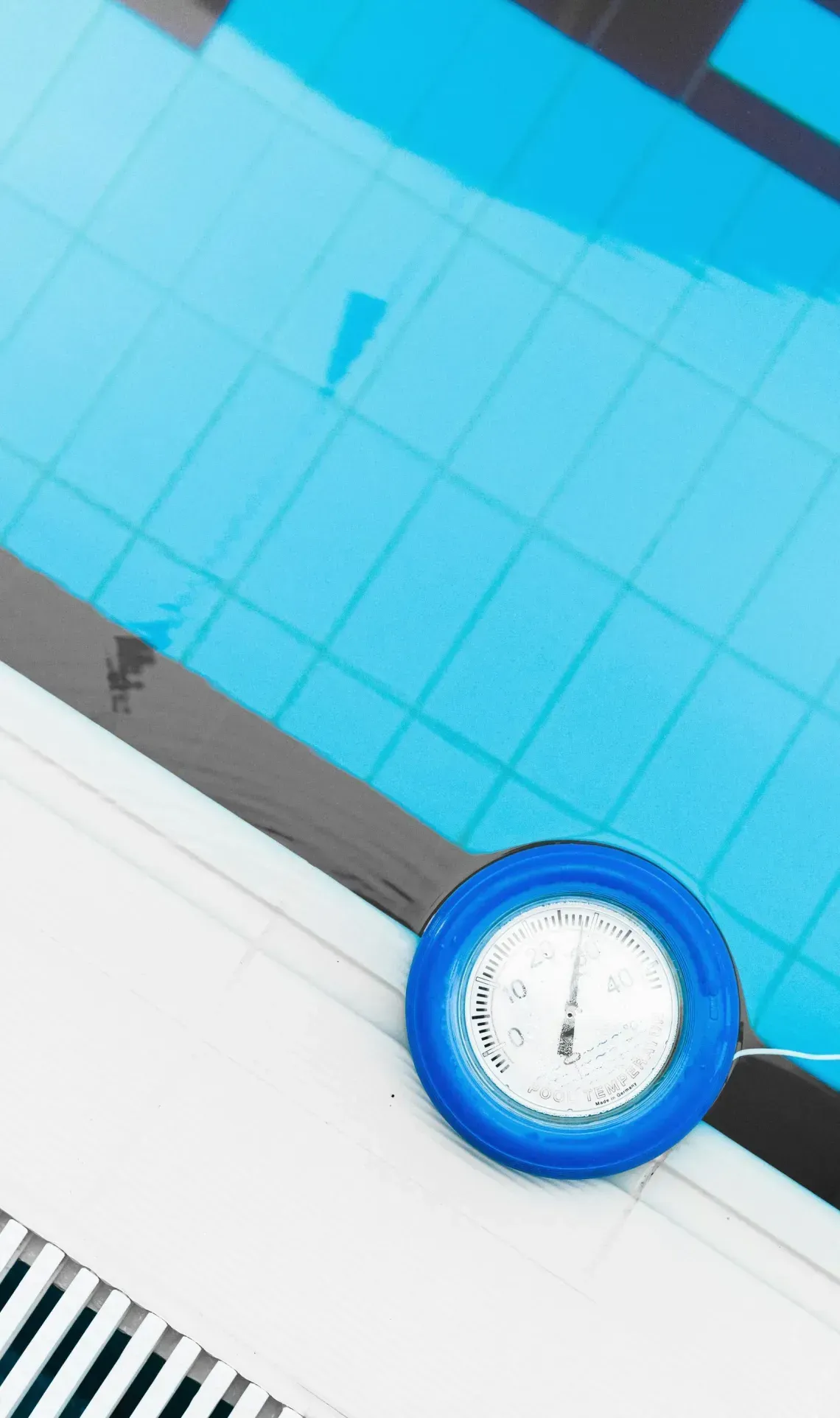A blue circular pool thermometer floats in clear blue water at the edge of a tiled swimming pool.