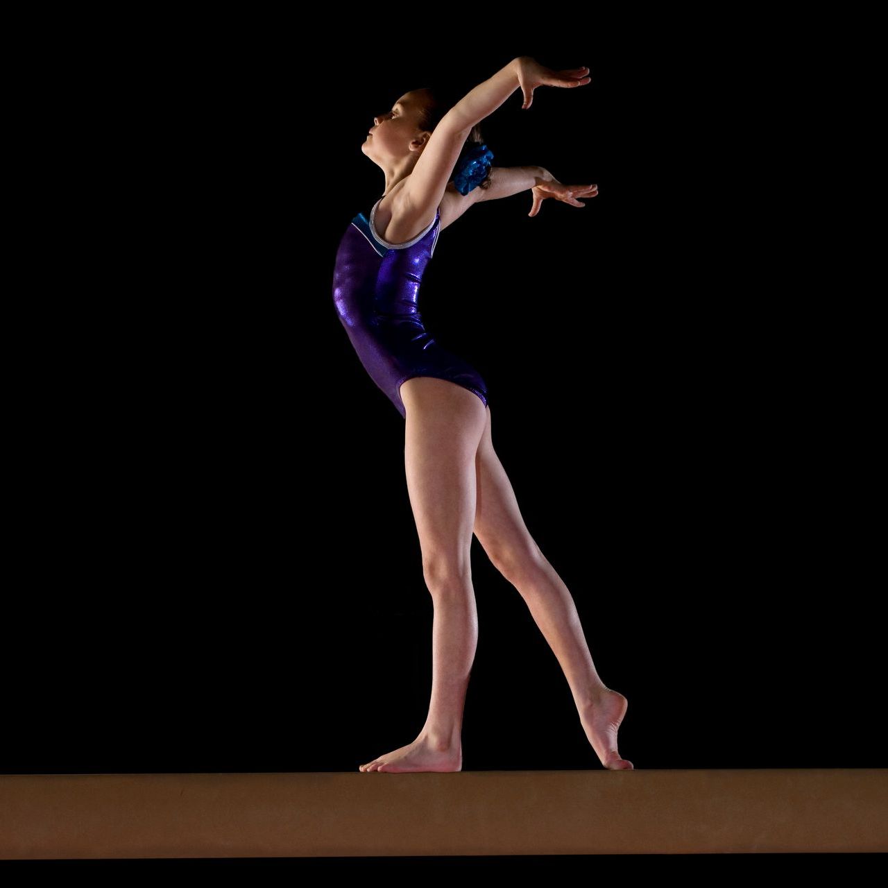 Gymnast in a purple leotard balances on a beam, arms raised, against a black background.