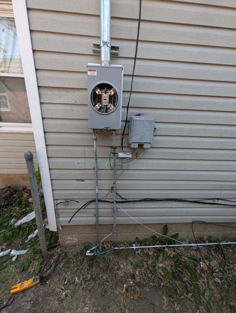 Exterior electrical meter box on a beige house with metal conduits and grounding wires visible.
