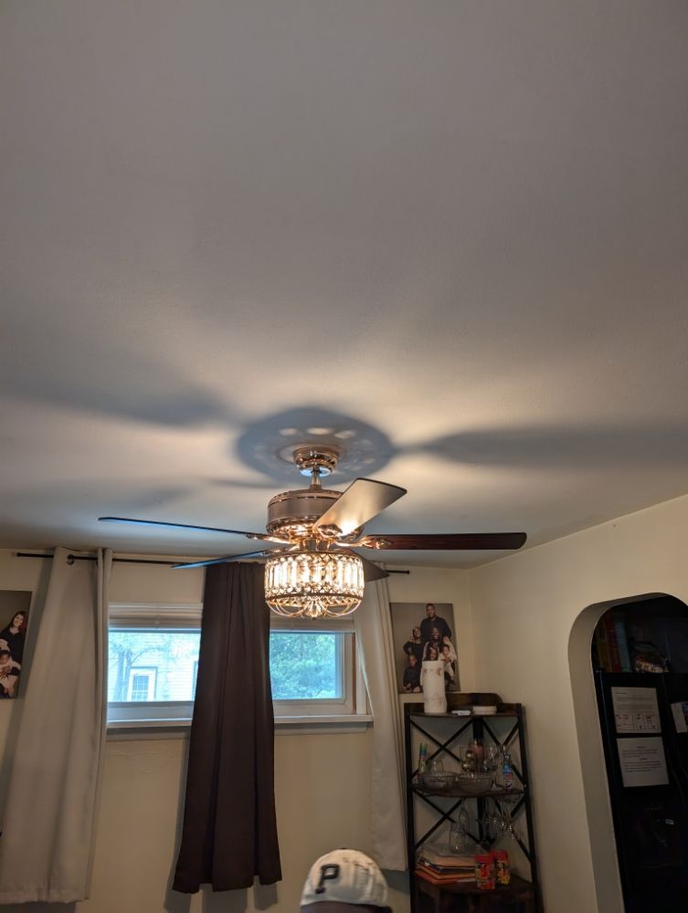 Ceiling fan with a decorative light fixture, casting shadows on the white ceiling; window in the background.