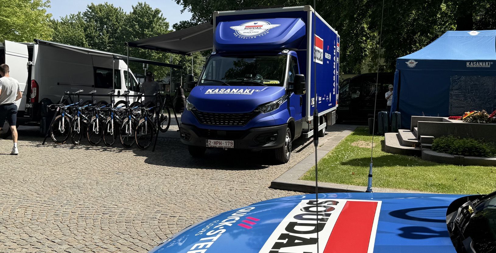 Blue van parked on gravel beside bicycles and tents at an outdoor event, with a banner in the foreground
