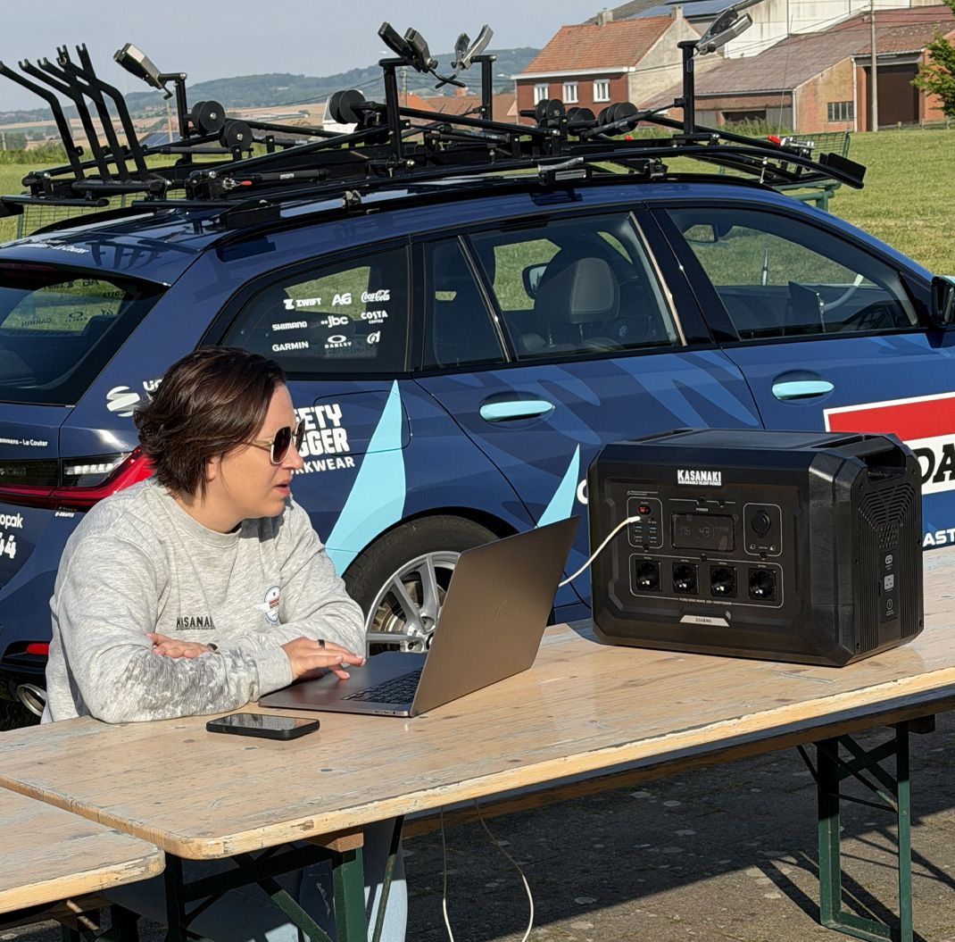 Person working on a laptop with Kasanaki K2 at a table beside a blue car with roof-mounted equipment outdoors.