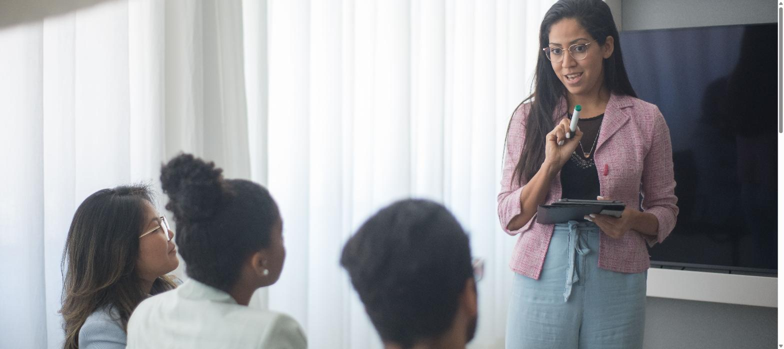 Woman in pink blazer presenting to colleagues in a bright meeting room