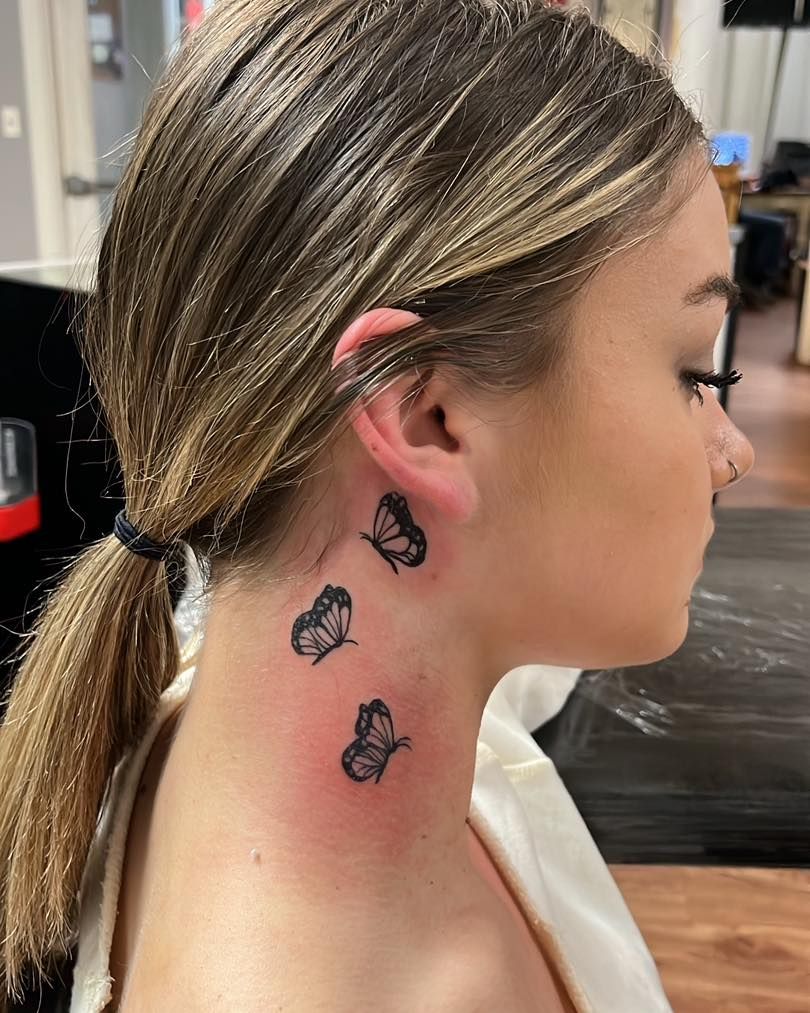 Woman with butterfly tattoos on neck, in a tattoo parlor. Blonde hair, ponytail. Black ink.