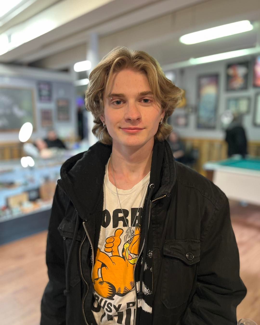 Young person with light hair and a black jacket smiles in a room with a pool table and art.