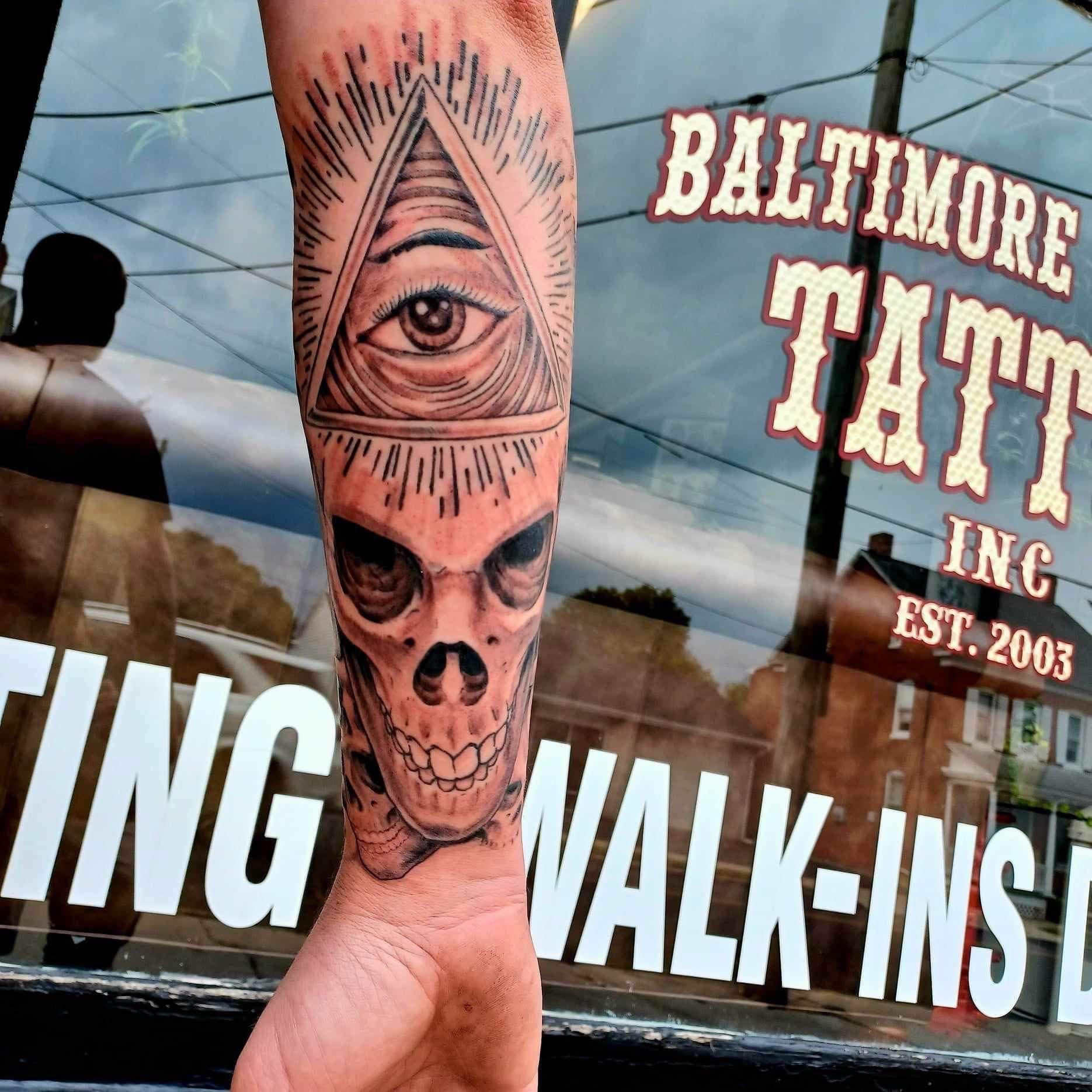 Tattoo of a skull and eye within a triangle on a forearm, outside a tattoo shop.
