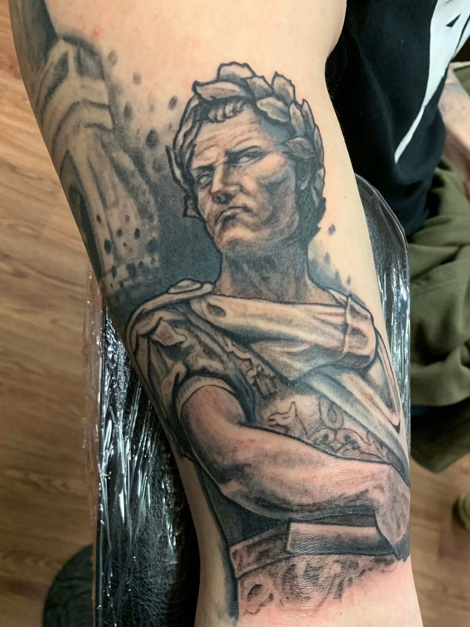 Tattoo of Julius Caesar with a laurel wreath, arms crossed. Black and grey shading.
