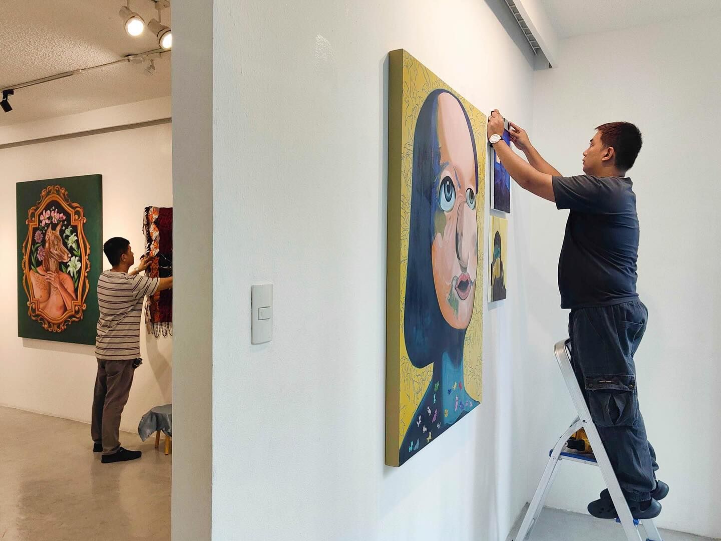 Two people hang artwork in a gallery. One stands on a ladder, the other adjusts a painting on the wall. 