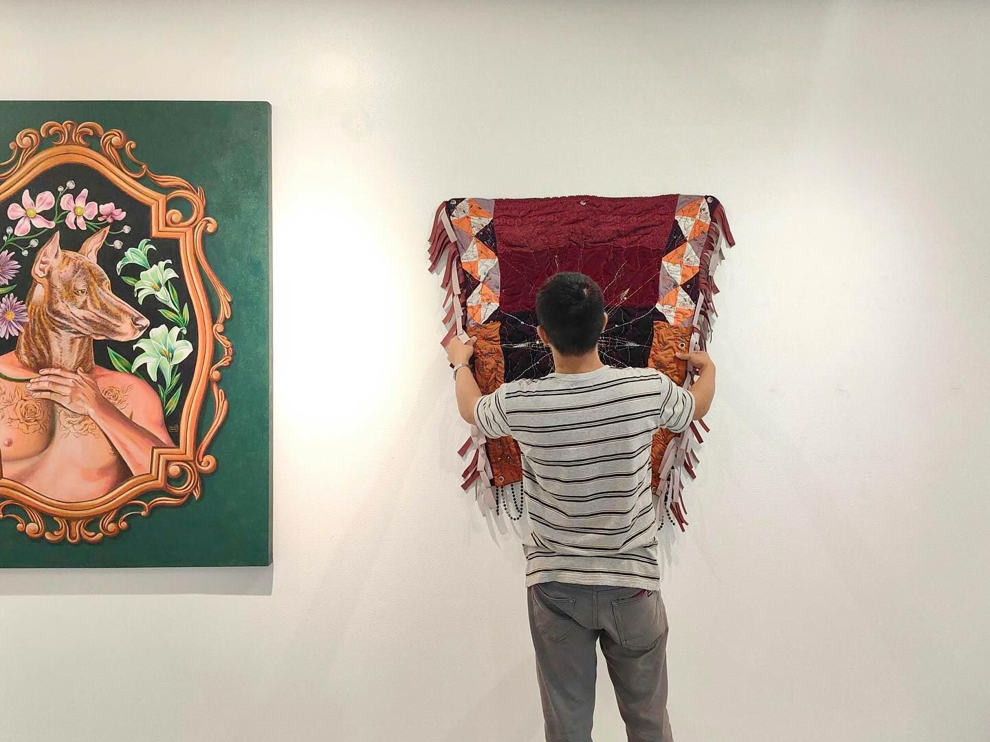 Person hangs a fringed textile on a white wall in a gallery next to a painting of a fox-like figure.