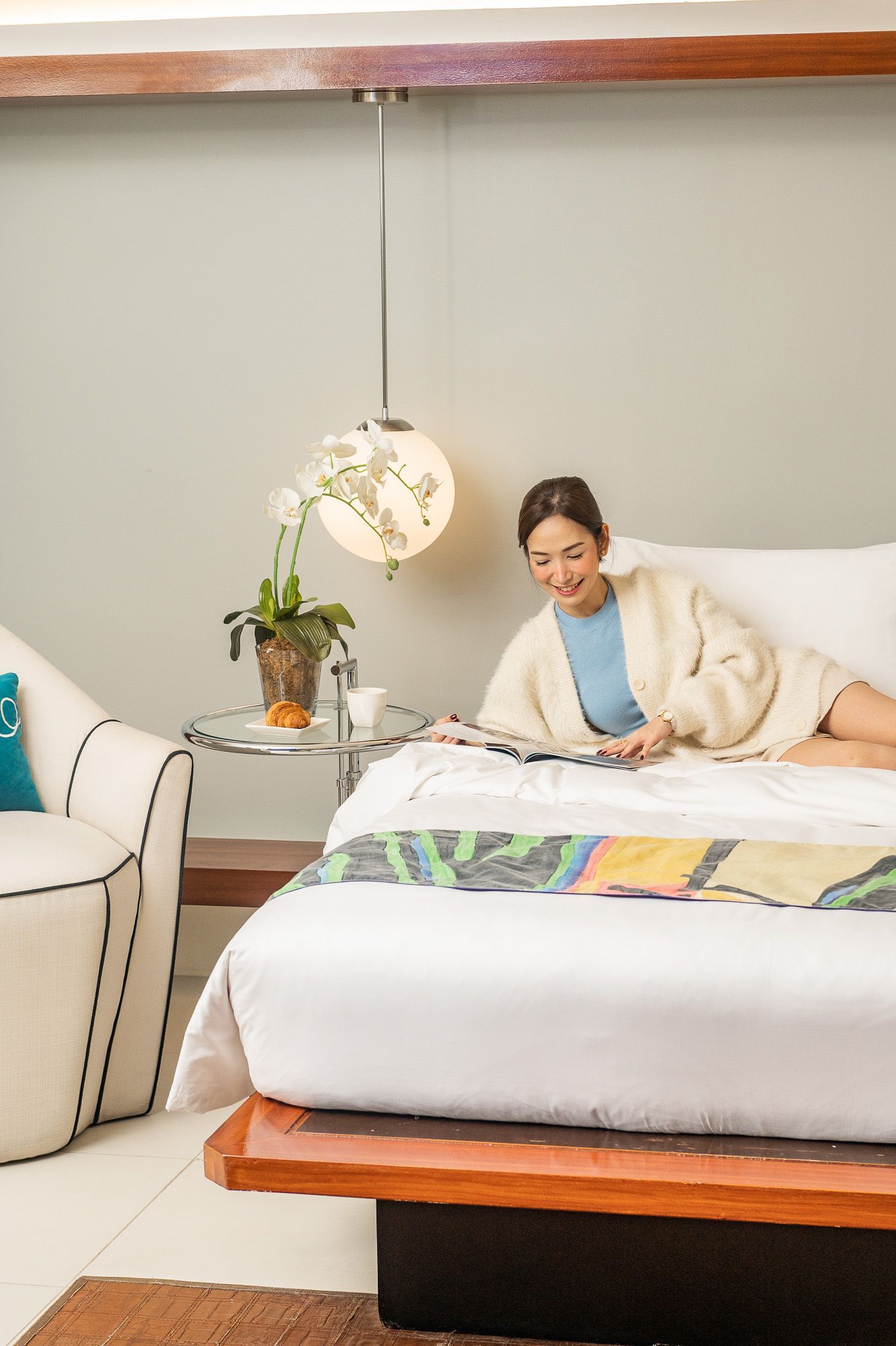 Woman relaxing on a white bed in a cozy bedroom, with a hanging lamp and side table nearby.