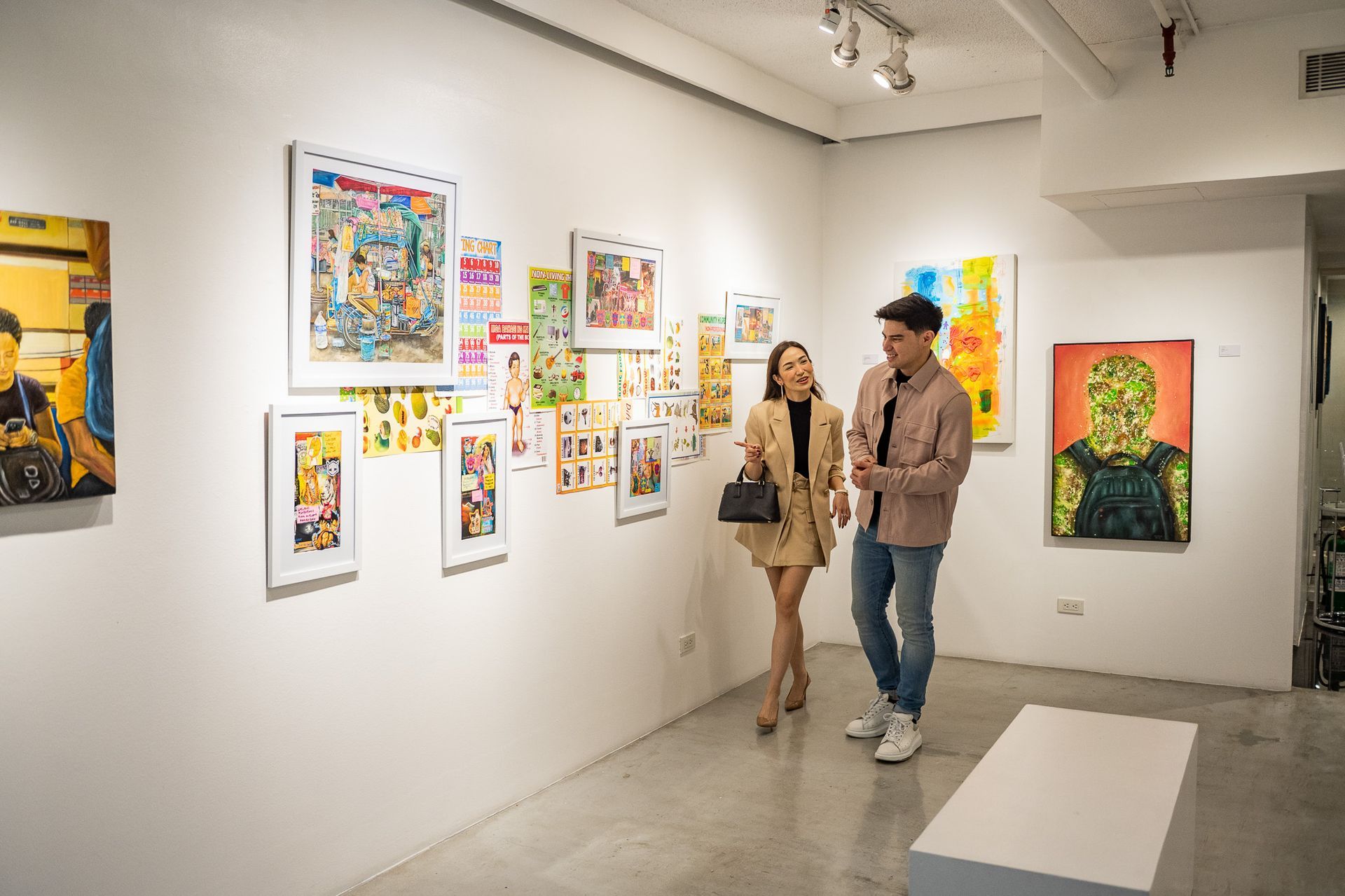Two people walking through a bright art gallery with colorful framed paintings on white walls