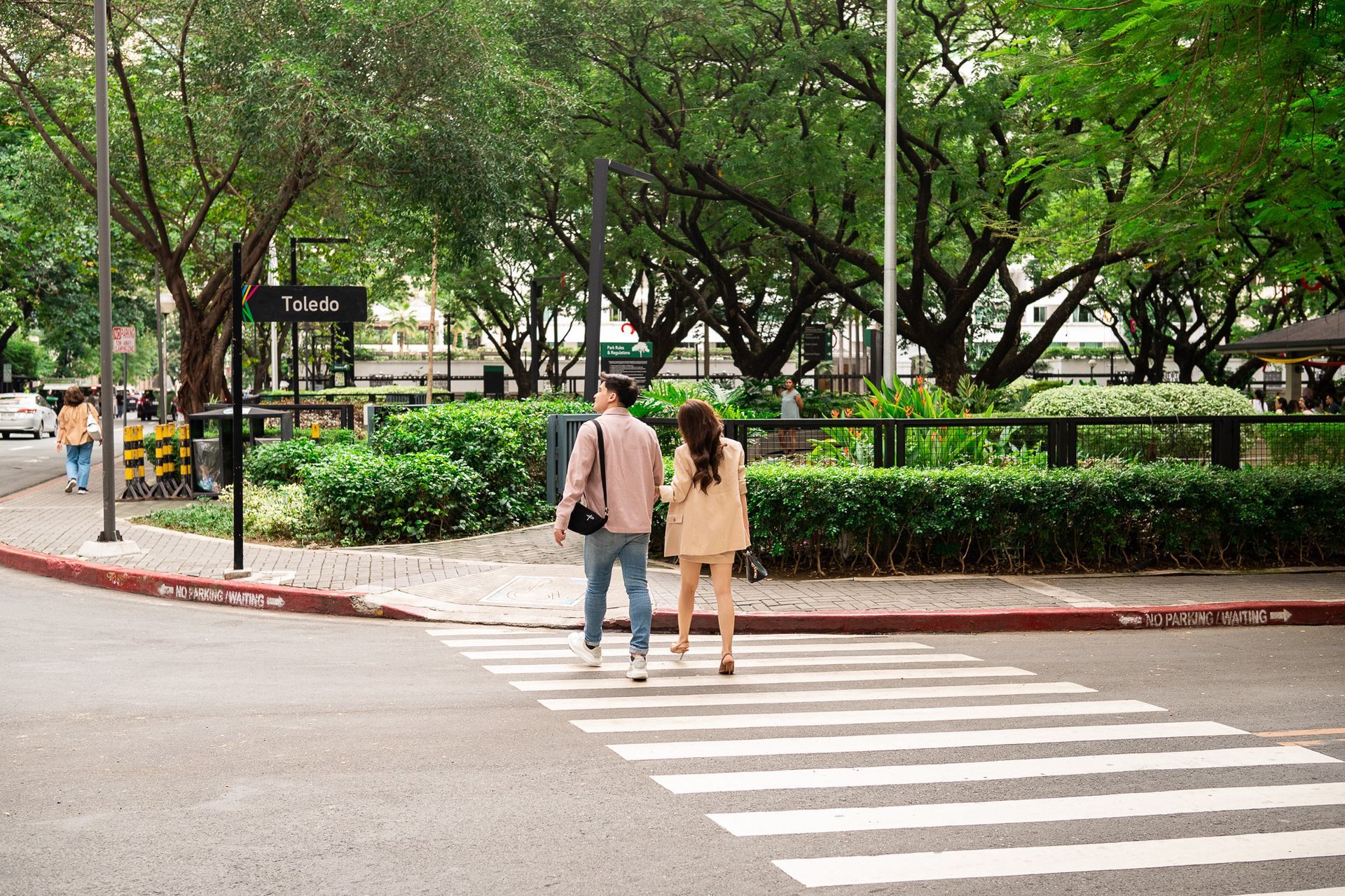 Two people crossing a street at a crosswalk in a leafy park with trees and a sign nearby