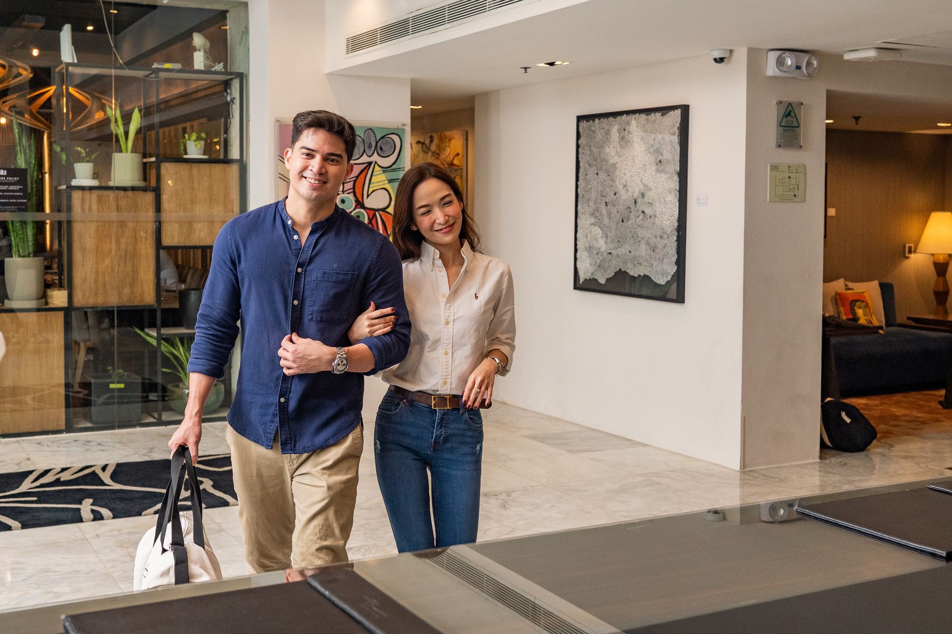 Two people walking through a modern lobby, smiling and carrying bags.