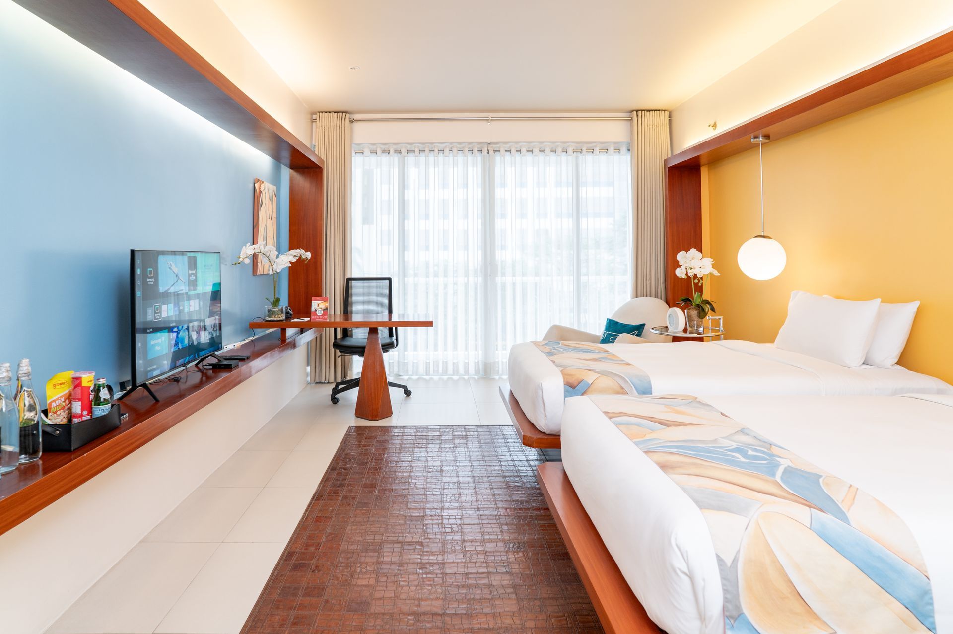 Bright modern hotel room with two beds, large window, desk, and colorful blue and orange walls