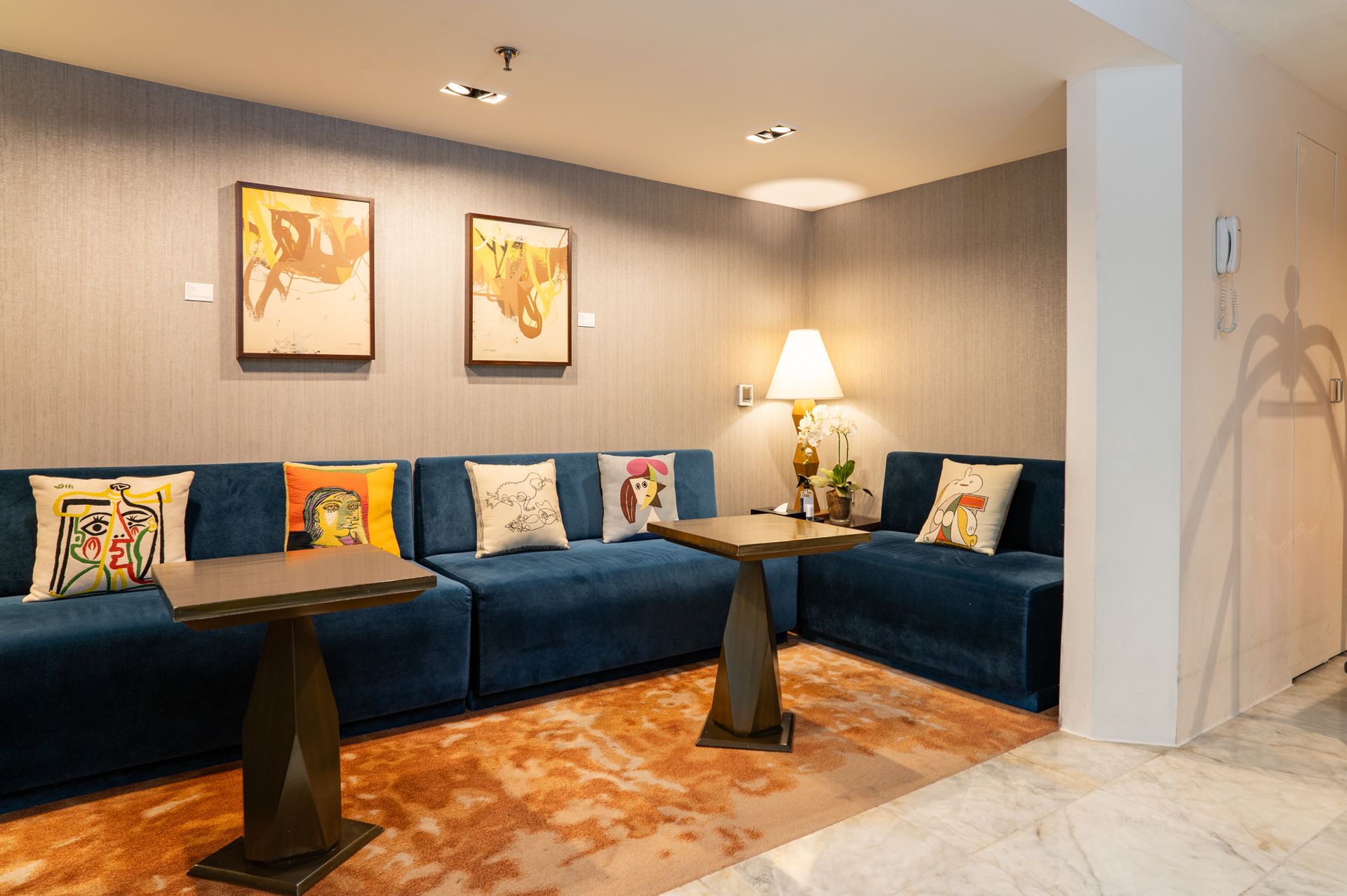 Cozy lounge area with blue velvet seating, art, lamps, and small tables on a patterned rug.