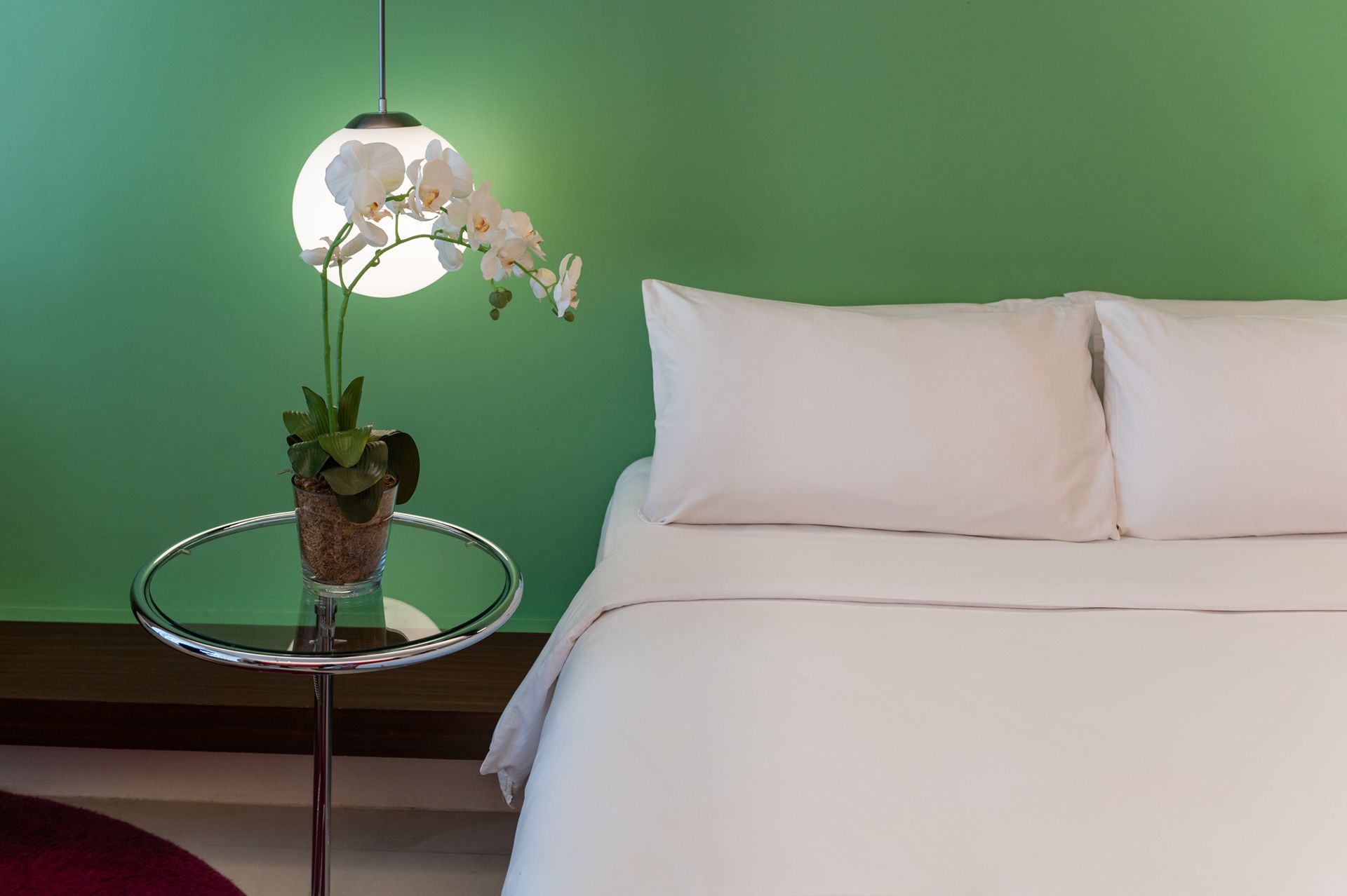 Bed with white linens, side table with orchid, under a pendant light against green wall.