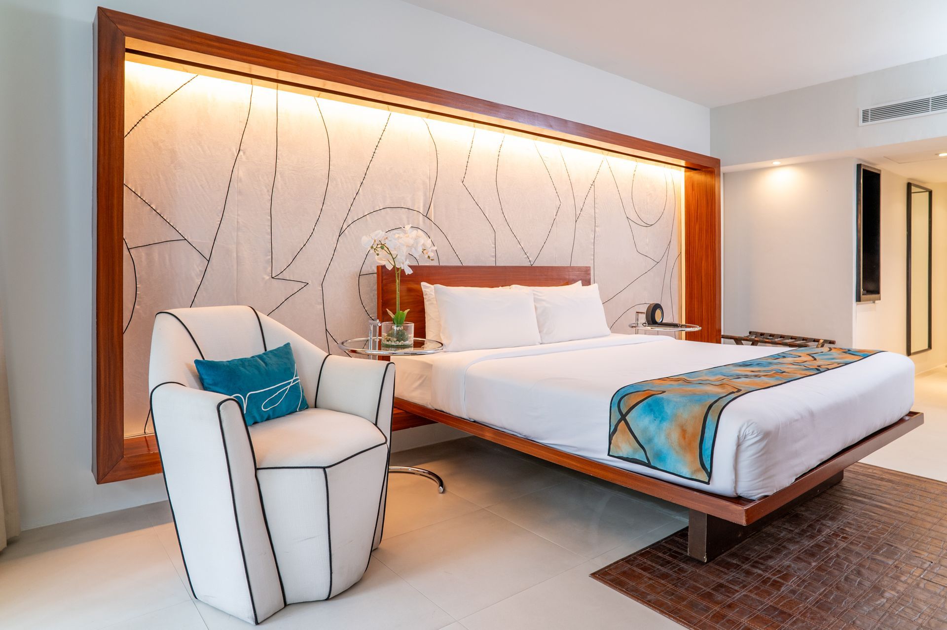 Hotel bedroom with a bed, white chair, and art on the wall. Brown, white, and blue colors.