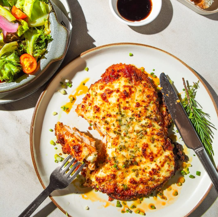 Chicken cutlet with cheese, fork in piece, side of salad, small bowl of sauce.