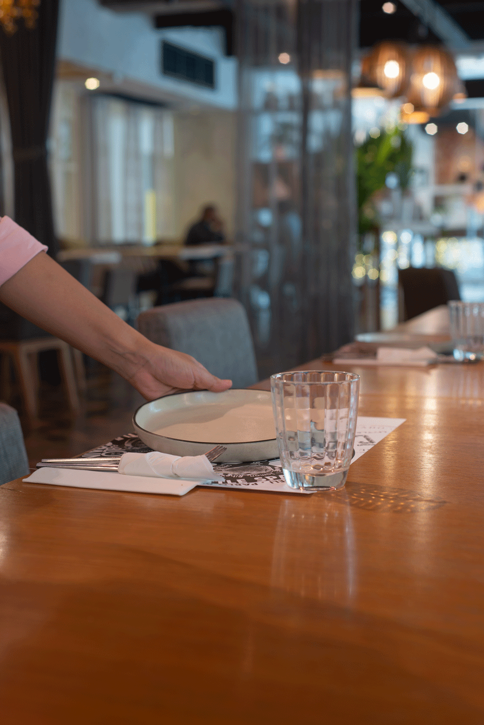 A hand placing a plate on a table set for dining, with a glass and paper placemat.