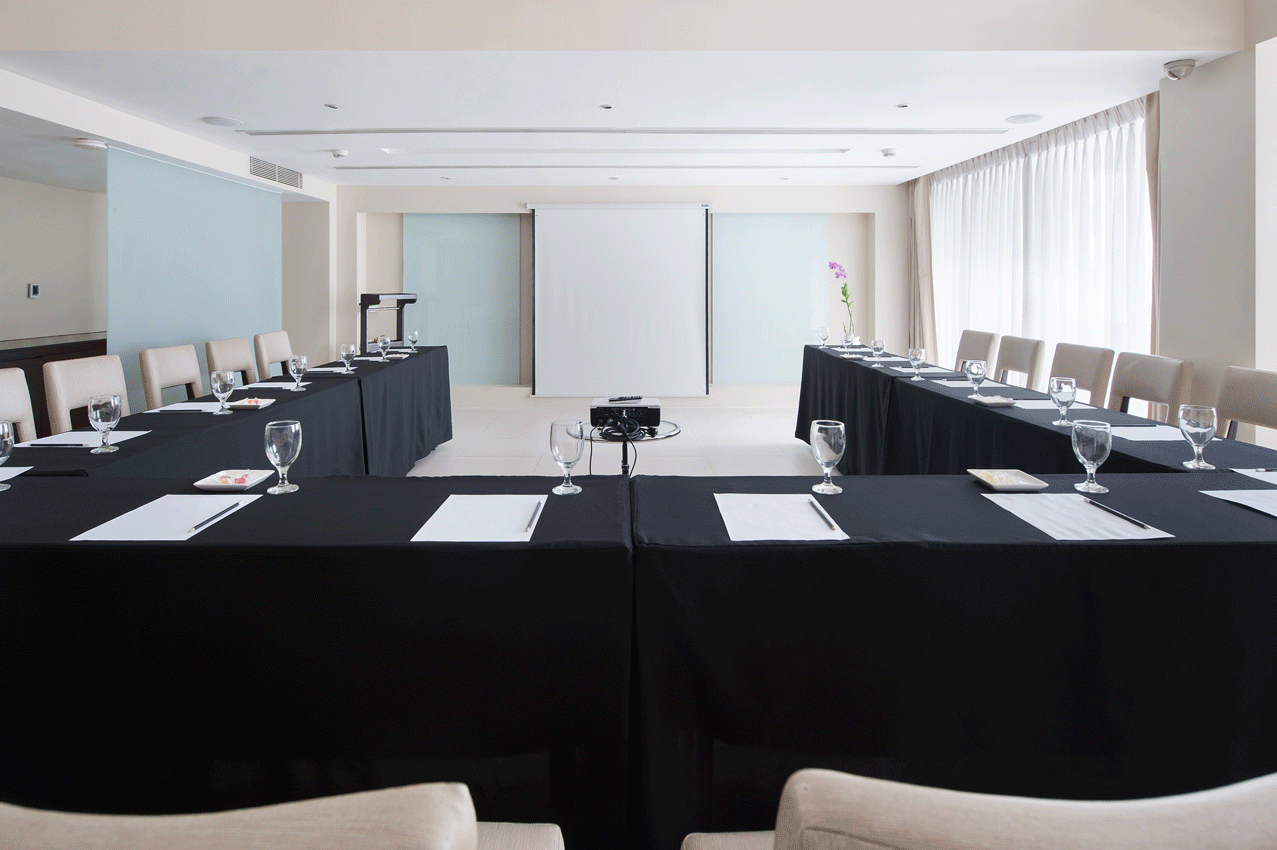 Conference room with U-shaped table setup, projector screen, white walls, and chairs.