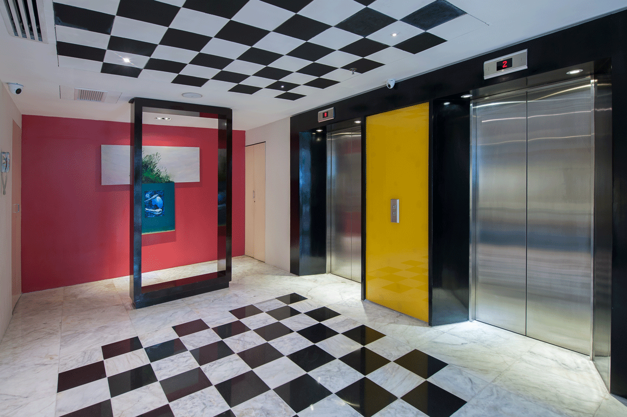 Elevator lobby with black, white, and yellow checkerboard pattern. Red wall with framed art. Stainless steel elevators.