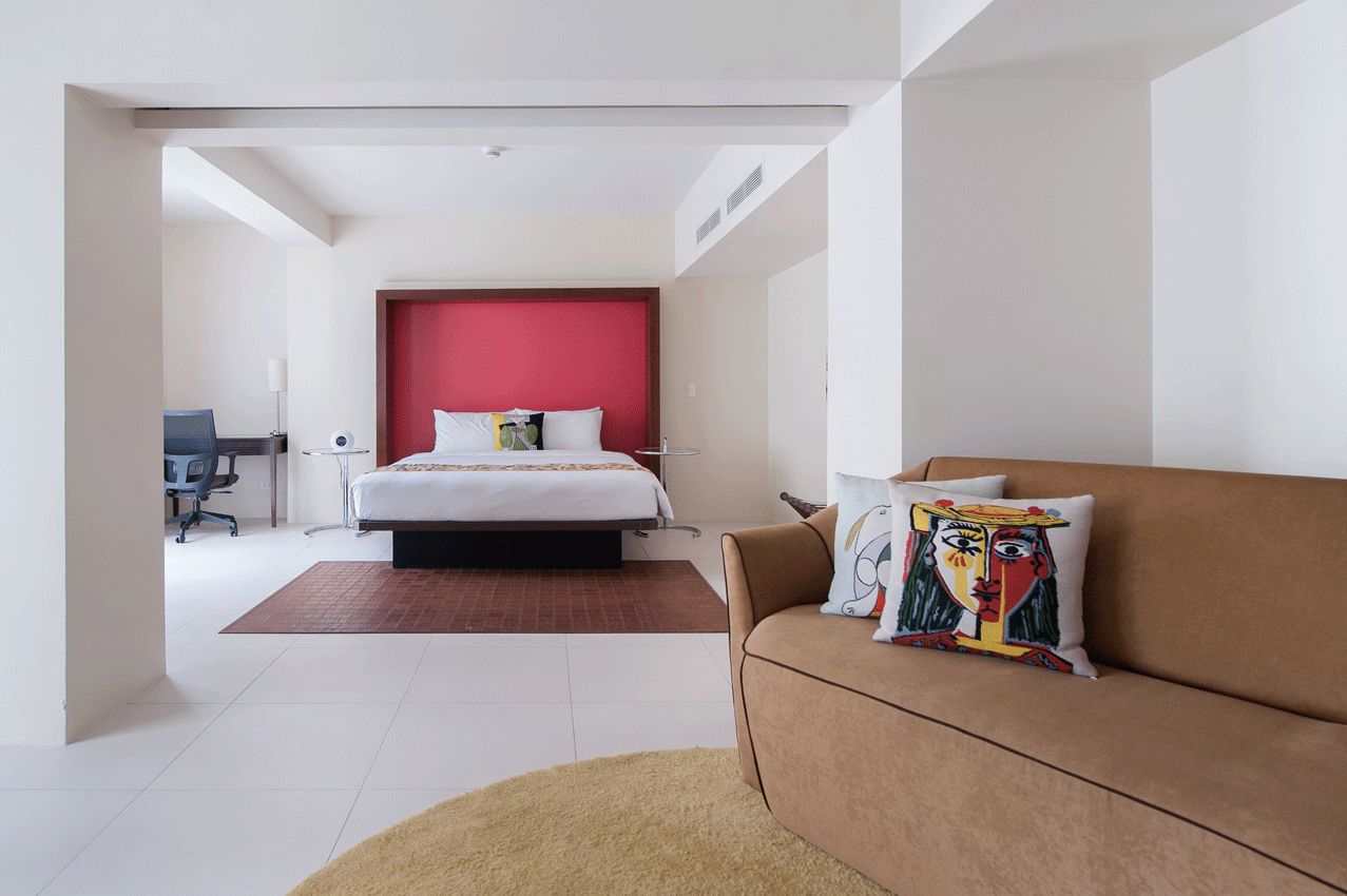 Hotel room with bed, sofa, desk, and area rugs; white walls, red headboard.