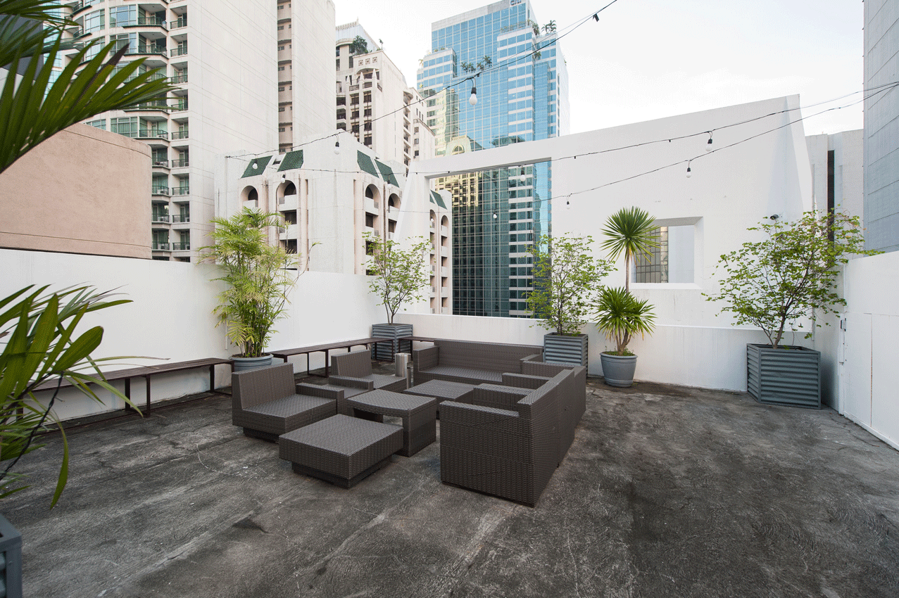 Rooftop lounge with gray wicker furniture, potted plants, and city buildings in background.