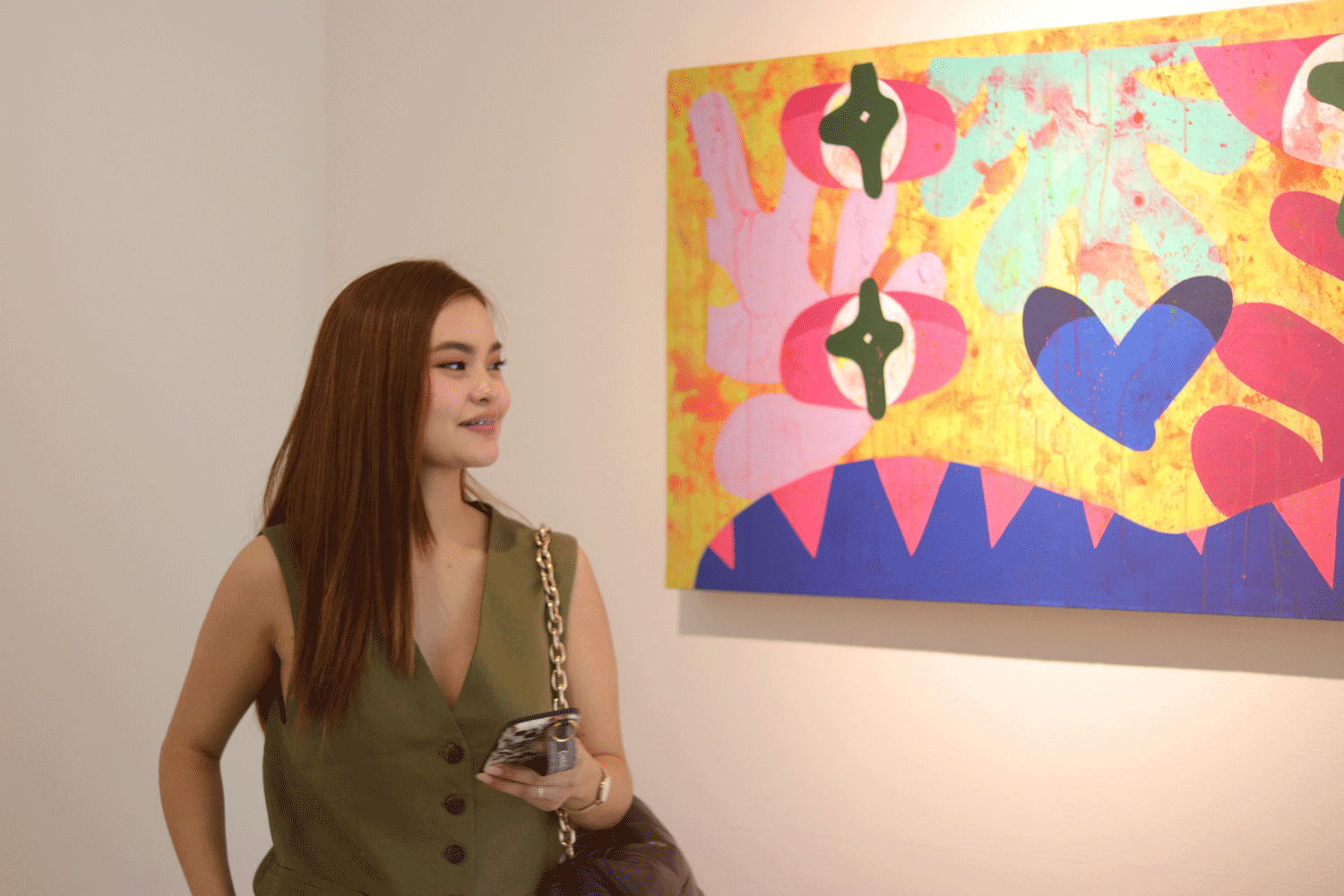 Woman viewing abstract art in a gallery; wearing green vest, holding phone, looking at the colorful painting.