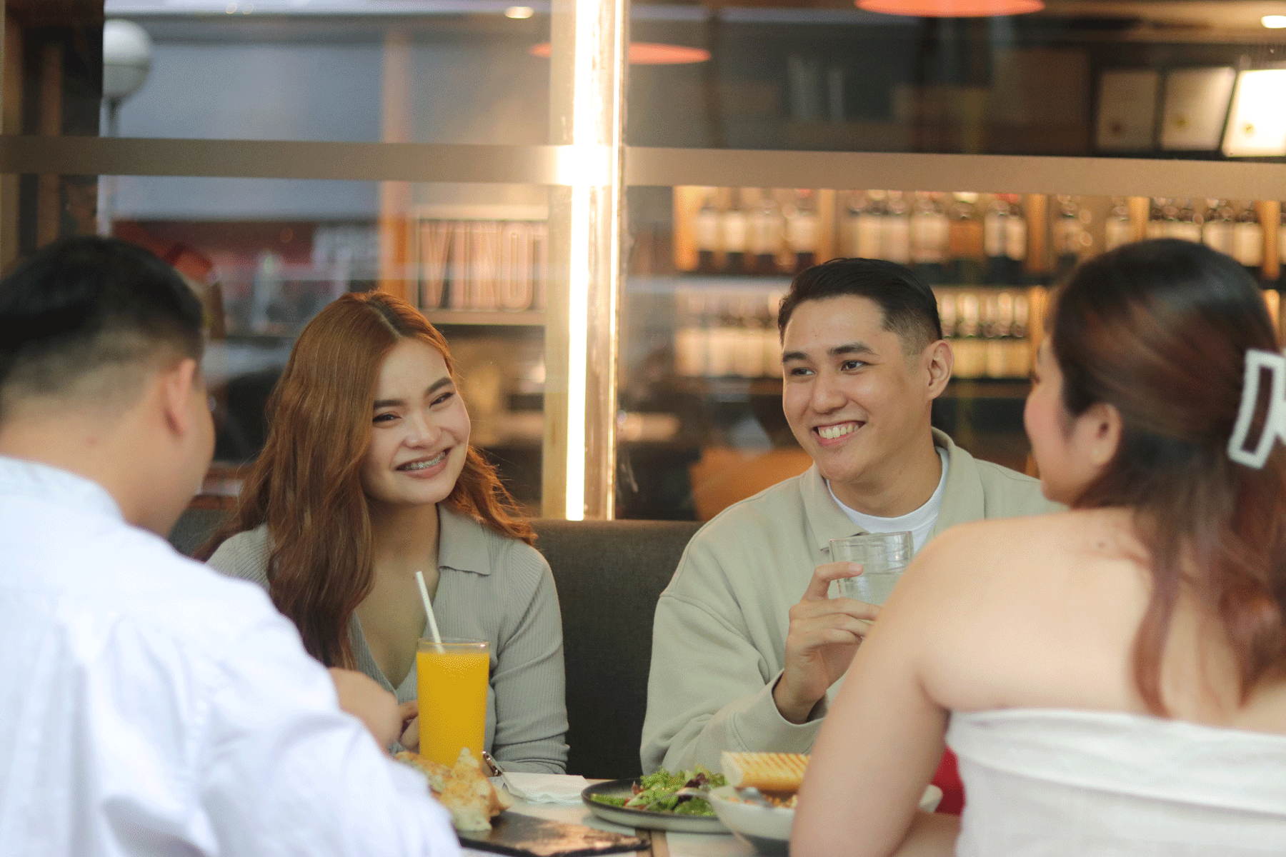 Friends chatting at a café table, smiling with drinks and food in a bright indoor setting.