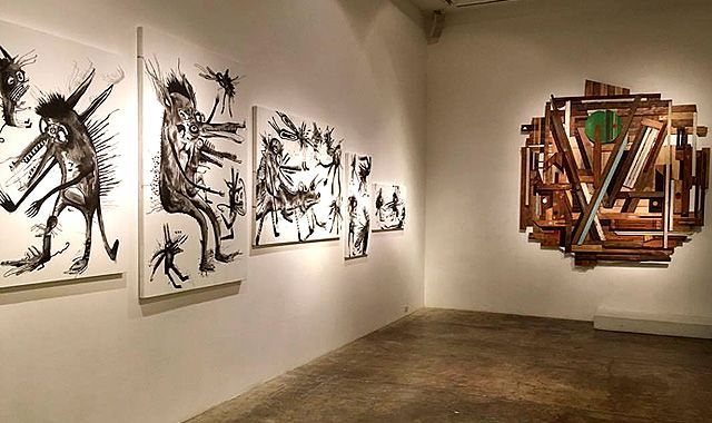 Art gallery interior with ink drawings of figures and a wooden wall sculpture.