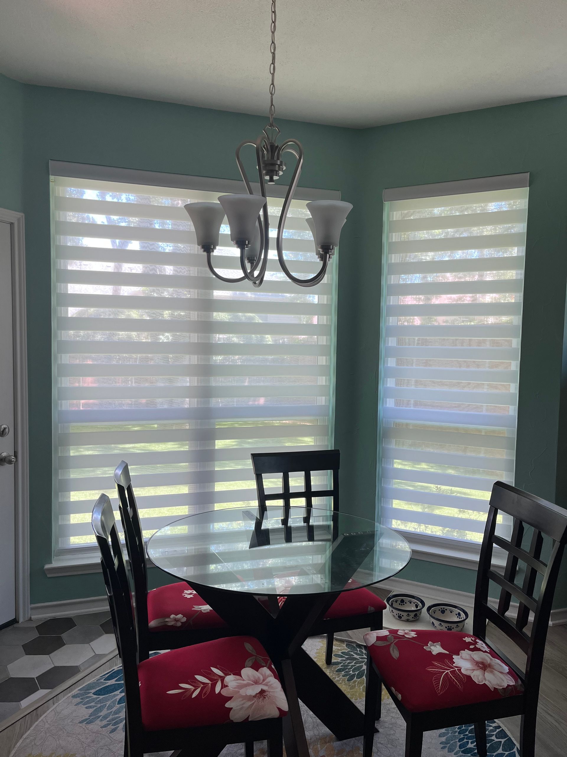 A dining room with round table, chairs with floral cushions, and window blinds; chandelier hangs above.