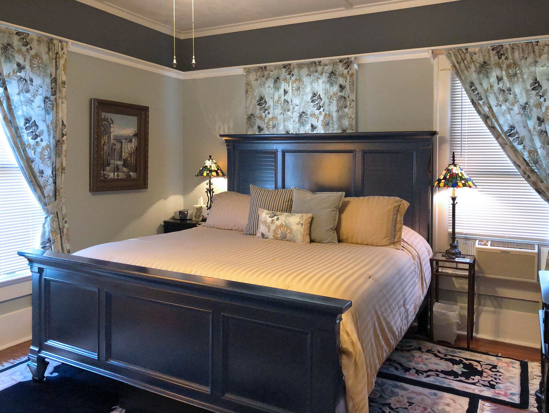 A bedroom with a large bed , two lamps , and two windows.