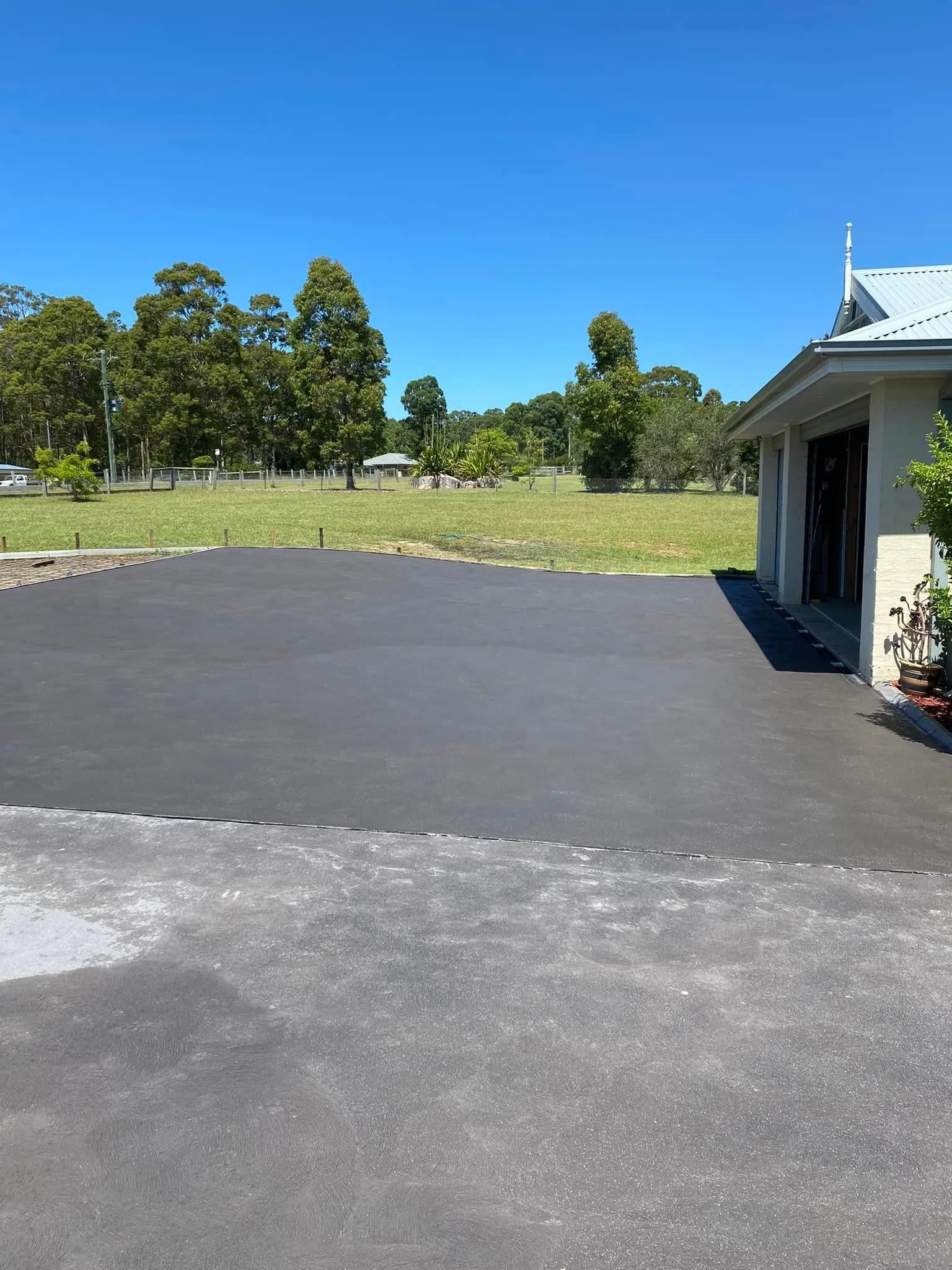 Empty driveway beside a garage, with a grassy yard, trees, and clear blue sky in the background — BJ's Concreting in Callala Bay, NSW