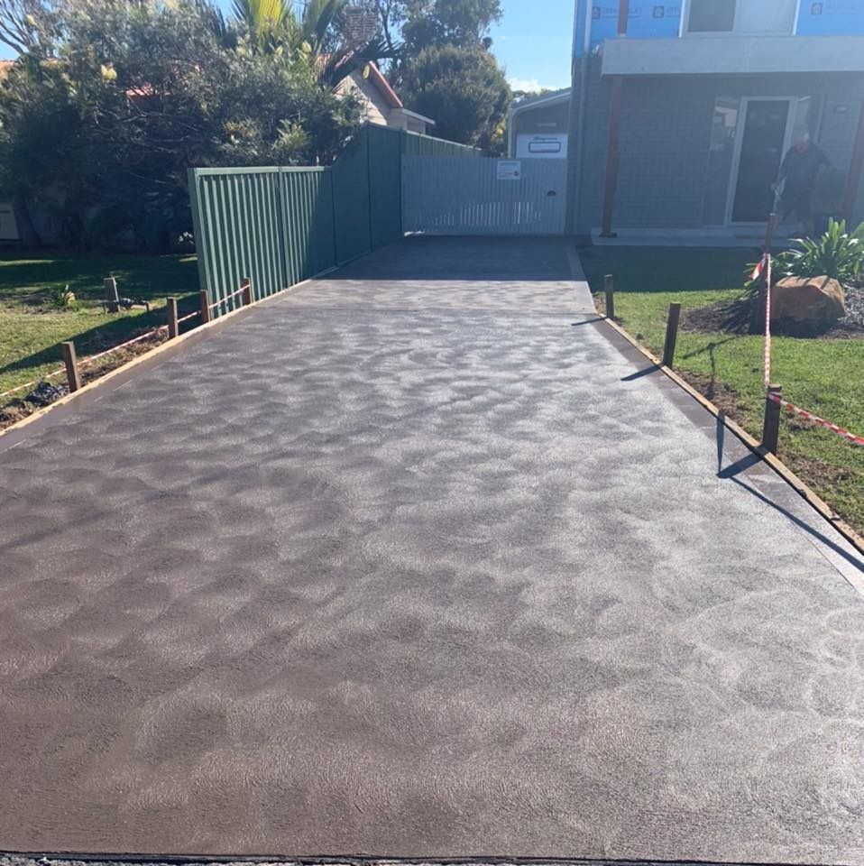 Freshly paved gray driveway beside a house and green fence in bright sunlight — BJ's Concreting in Bomaderry, NSW