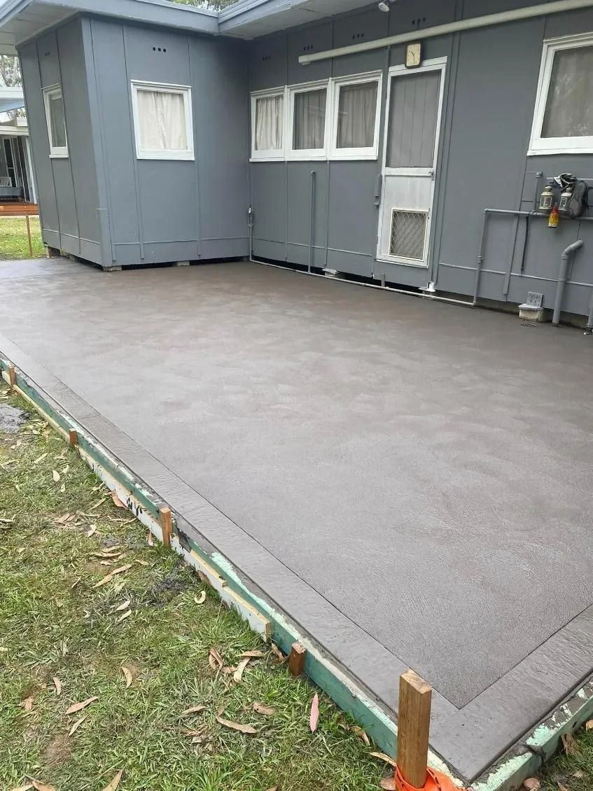 Freshly poured concrete patio beside a gray house with windows and doors, bordered by grass and small stakes — BJ's Concreting in Bomaderry, NSW